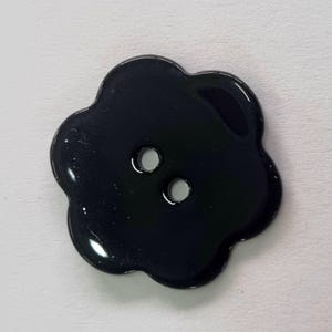 Puede incluir: Un botón negro en forma de flor con dos agujeros para coser. El botón tiene un acabado brillante y un borde festoneado. Este botón decorativo es adecuado para varios proyectos de costura.