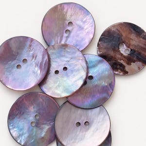 Puede incluir: Un conjunto de siete botones redondos de dos agujeros con un acabado iridiscente y nacarado. Los botones muestran un espectro de colores que incluye tonos violeta intenso, azul y marrón con una superficie lisa y reflectante y textura de concha.