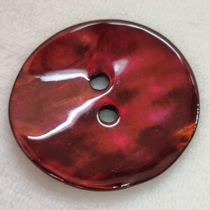 Könnte beinhalten: Ein großer, runder Knopf mit glänzend rotbraunem Finish. Der Knopf hat zwei Löcher und einen wirbelnden, marmorierten Effekt mit rosa und orangefarbenen Akzenten. Der Knopf ist wahrscheinlich aus Kunststoff oder Harz.