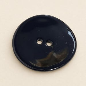 Op de afbeelding: Een ronde, glanzend zwarte knoop met een glad, reflecterend oppervlak en twee centrale gaten voor bevestiging. De knoop heeft een eenvoudige, afgeronde rand en een egale, donkere afwerking die het licht op het oppervlak weerkaatst.