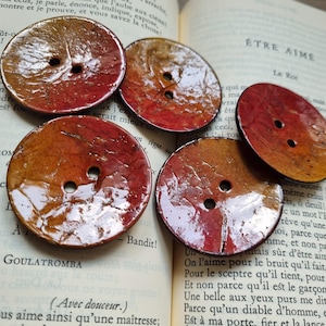 Puede incluir: Cuatro botones redondos de dos agujeros con un acabado brillante. Los botones están pintados en tonos de rojo, naranja y amarillo, creando un efecto degradado. Los botones están dispuestos sobre un libro con texto en francés.