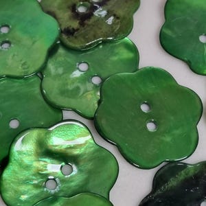 Op de afbeelding: Close-up van verschillende groene, bloemvormige knopen. Elke knoop heeft twee gaten en een glanzende, iriserende afwerking. De groentinten variëren, waarbij sommige knopen donkerder, bijna zwart lijken.