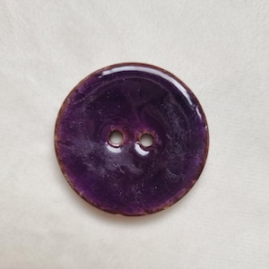 May include: A large, circular button with two holes. The button is a deep purple colour with a glossy finish and a brown edge. Suitable for sewing projects, the button is likely made of a durable material.