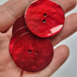 Può includere: Una collezione di grandi bottoni rotondi rossi con due fori ciascuno. I bottoni hanno una finitura lucida e iridescente, che riflette la luce. I bottoni sono probabilmente fatti di un materiale a conchiglia e sono adatti per progetti di cucito.