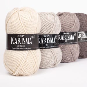 Op de afbeelding: Vier strengen Drops Karisma-garen op een rij, elk met een zwart etiket. De garenkleuren variëren van crème tot verschillende tinten grijs en bruin. De etiketten lezen "DROPS KARISMA" en "100% Wool".