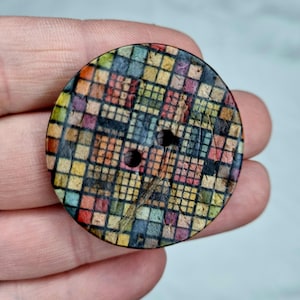 Puede incluir: Un botón redondo de madera con un patrón geométrico colorido. El botón tiene dos agujeros y mide aproximadamente 2,5 cm de diámetro.