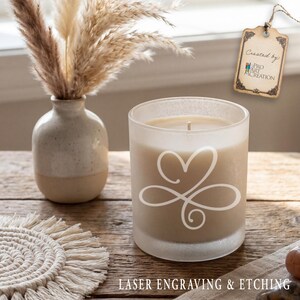 Puede incluir: Una vela de vidrio esmerilado con un dise&ntilde;o de coraz&oacute;n y espiral, una mecha encendida y un relleno de cera beige. La vela est&aacute; sobre una superficie de madera junto a un peque&ntilde;o jarr&oacute;n con plantas secas y un posavasos de macram&eacute;. El texto en la parte inferior dice "LASER ENGRAVING & ETCHING."