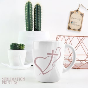 May include: White ceramic mug featuring a pink heart, cross, and dove design. The mug has a curved handle and is surrounded by white planters with cacti and succulents. A decorative tag hangs in the background.