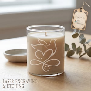 Peut inclure: Une bougie beige dans un pot en verre transparent, avec un motif blanc grav&eacute; d'une colombe et d'un c&oelig;ur. La bougie est pos&eacute;e sur une surface en bois, avec une petite coupelle et une &eacute;tiquette "Created by: Pro Art Creation". Le texte "LASER ENGRAVING & ETCHING" est en bas.