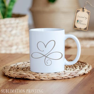 Puede incluir: Taza de cer&aacute;mica blanca con un dise&ntilde;o de coraz&oacute;n y remolino gris. La taza est&aacute; sobre un posavasos tejido. Las palabras "Sublimation Printing" est&aacute;n en la parte inferior de la imagen. Una etiqueta que dice "Created by Pro Art Creation" cuelga en el fondo.