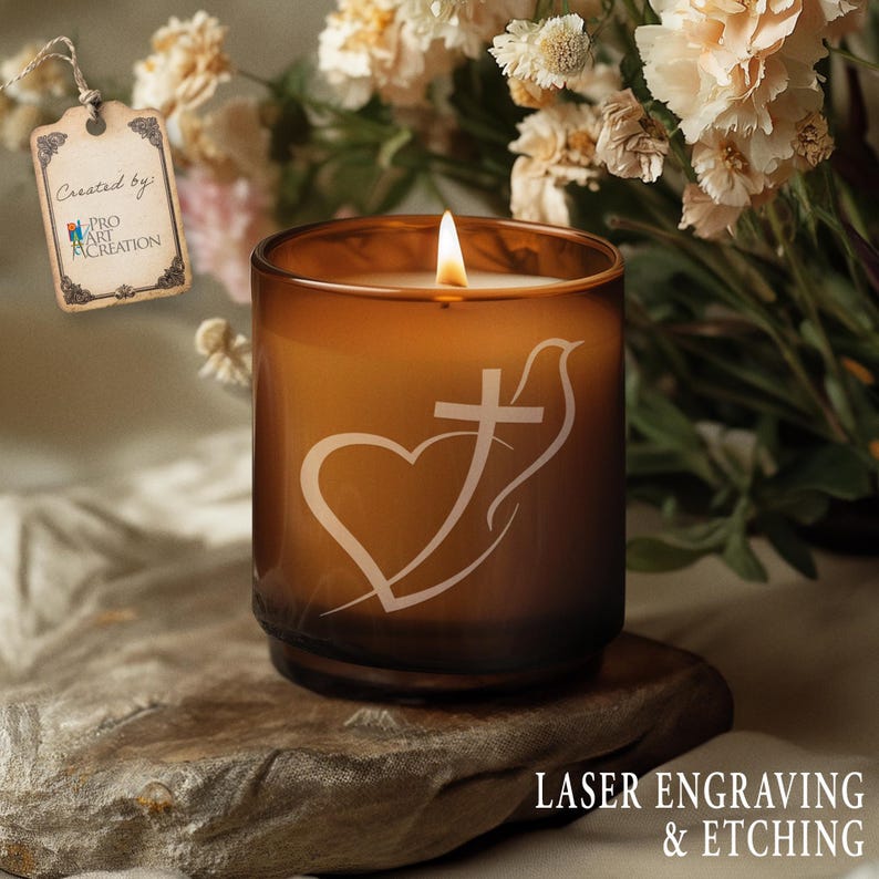 May include: A lit amber-colored candle in a glass jar with a white etched design of a heart, cross, and dove. The candle sits on a stone surface. The text "LASER ENGRAVING & ETCHING" is visible.