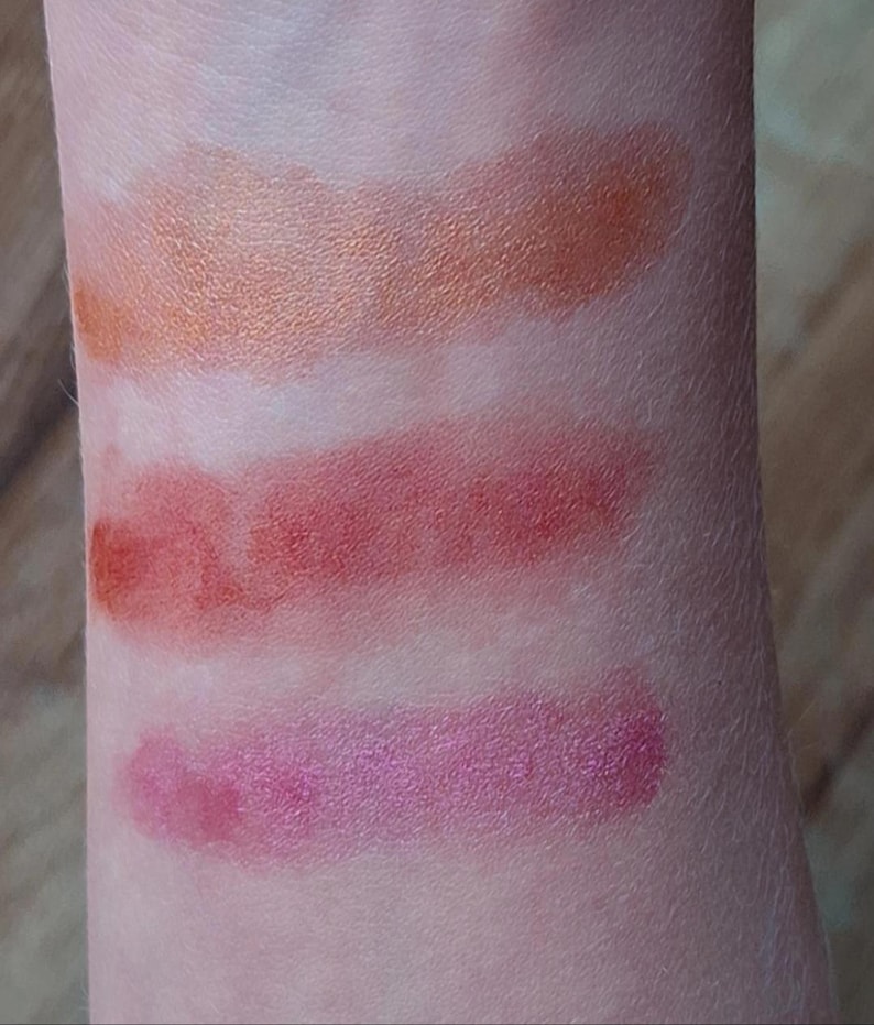 Puede incluir: Primer plano de un brazo con cuatro muestras de maquillaje. Las dos superiores son de color naranja y bronce, las dos inferiores son de color rosa y rojo. El maquillaje est&aacute; aplicado sobre la piel, mostrando los colores y texturas.