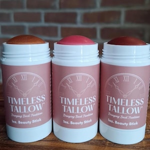 Puede incluir: Tres barras de belleza en envases blancos con etiquetas rosas. Las etiquetas dicen "TIMELESS TALLOW" y "1 oz. Beauty Stick". Las barras tienen diferentes tonos de color. El fondo es una pared de ladrillos.