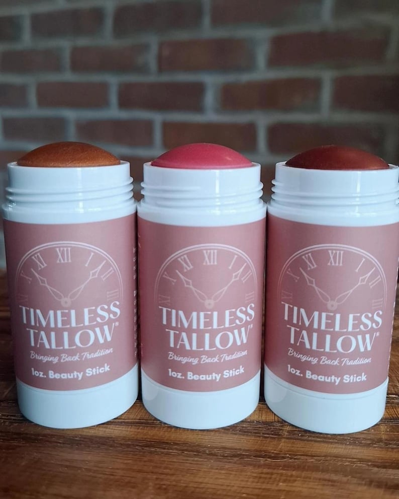 Puede incluir: Tres barras de belleza en envases blancos con etiquetas rosas. Las etiquetas dicen "TIMELESS TALLOW" y "1 oz. Beauty Stick". Las barras tienen diferentes tonos de color. El fondo es una pared de ladrillos.