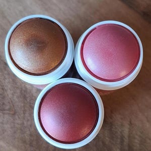 Puede incluir: Tres polveras de maquillaje con acabados met&aacute;licos. Una es bronce, otra rosa y la tercera roja intensa. Cada polvera tiene un borde blanco y una tapa met&aacute;lica. Los productos est&aacute;n dispuestos sobre una superficie de madera.