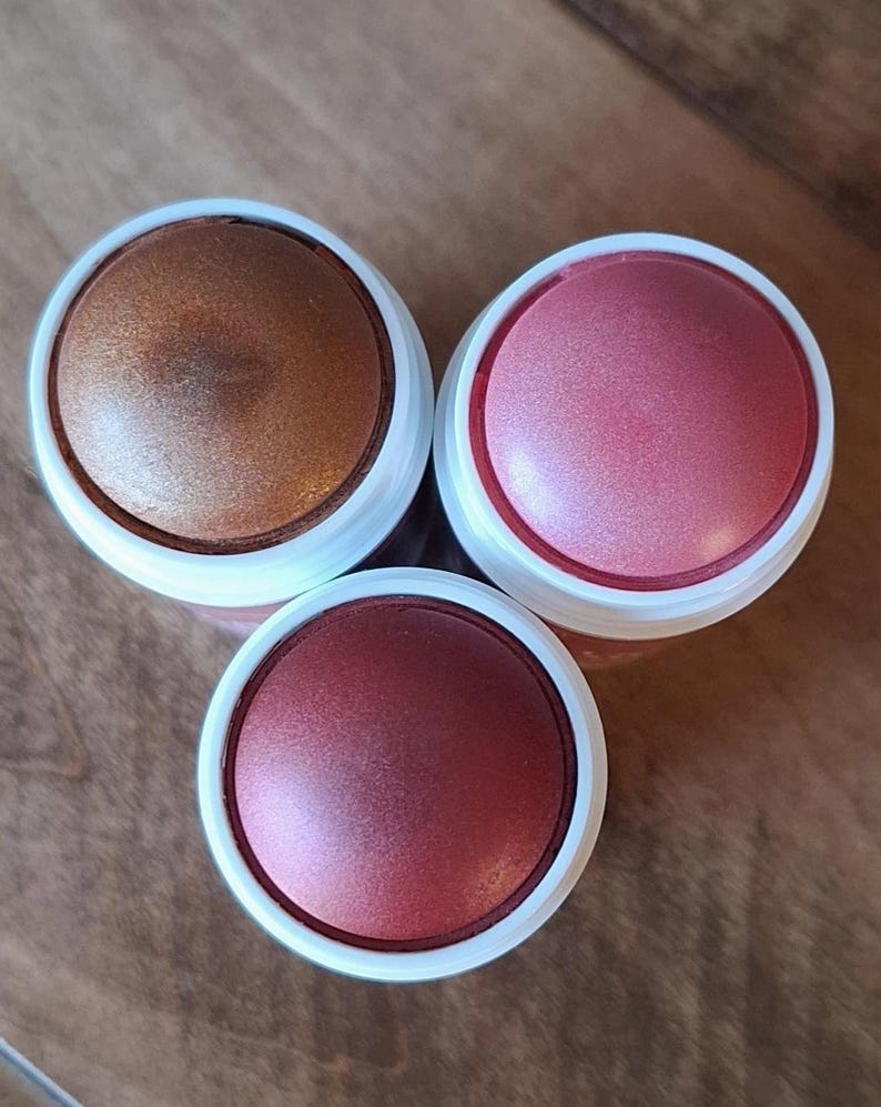 Puede incluir: Tres polveras de maquillaje con acabados met&aacute;licos. Una es bronce, otra rosa y la tercera roja intensa. Cada polvera tiene un borde blanco y una tapa met&aacute;lica. Los productos est&aacute;n dispuestos sobre una superficie de madera.