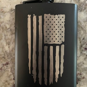 May include: A black stainless steel flask with a brushed silver American flag design. The flask has a hinged cap and a matte finish. The flag design features stars and stripes, giving it a patriotic theme.