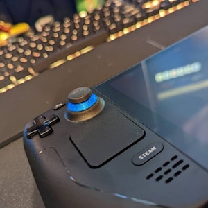 May include: A black handheld gaming device with a blue and black button, a black cross-shaped button, and a "STEAM" button. The device has a black screen and a speaker grill.