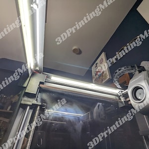 May include: A close-up view of a 3D printer with a black and silver body. The printer is equipped with two white LED lights. The printer is in a room with a blue wall and a white ceiling.