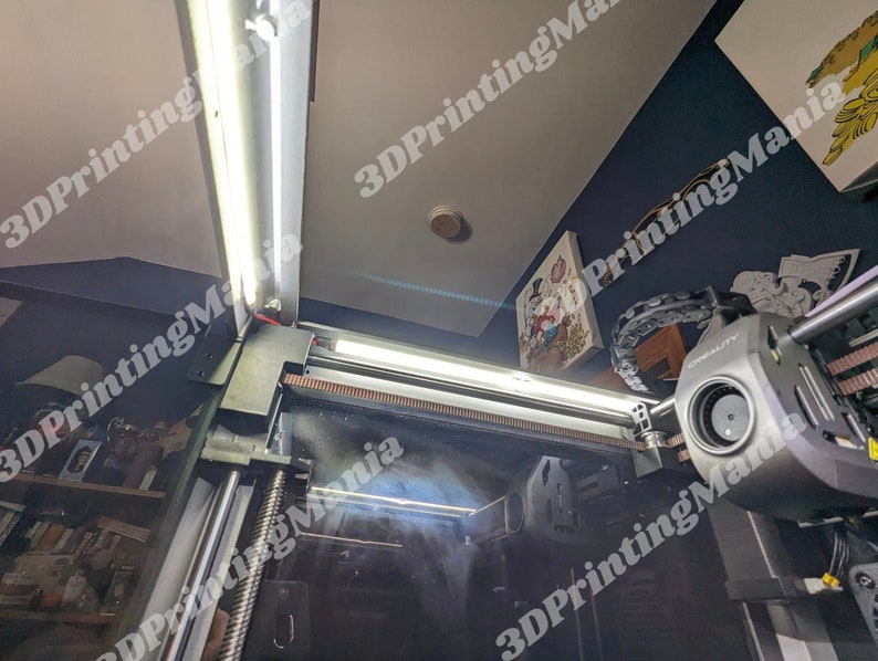 May include: A close-up view of a 3D printer with a black and silver body. The printer is equipped with two white LED lights. The printer is in a room with a blue wall and a white ceiling.