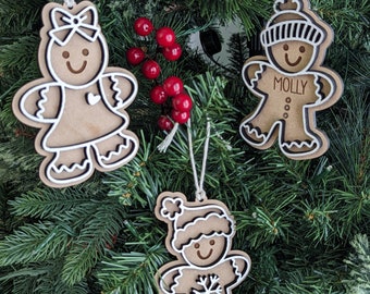 May include: Three wooden gingerbread person ornaments with white outlines. The ornaments are hanging on a green Christmas tree. One ornament has the name "Molly" written on it. Another ornament has a snowflake design. The third ornament has a bow.