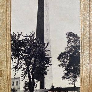 May include: Black and white vintage postcard featuring a tall, narrow, stone monument with a tree on either side. The text on the postcard reads "BUNKER HILL BATTLE MONUMENT, CHARLESTOWN, MASSACHUSETTS."