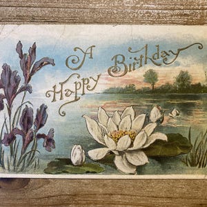 May include: Vintage postcard with a watercolor illustration. The card features a lake scene with a white water lily, purple irises, and the words "A Happy Birthday" in gold script. The background shows a sunset.