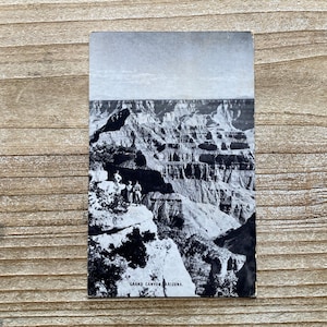 May include: Black and white vintage postcard featuring a scenic view of the Grand Canyon in Arizona. The postcard shows the canyon's layered rock formations and a group of people standing on a cliff. The text "Grand Canyon, Arizona" is printed at the bottom.