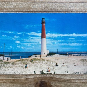May include: A vintage postcard depicting a red and white lighthouse situated on a sandy beach, with a blue sky and white clouds. People can be seen walking on the beach in the distance.