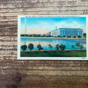 May include: A vintage postcard featuring a color illustration of the Bureau of Engraving and Printing and the Washington Monument near the Potomac Park Basin in Washington, D.C., with the text M516-NEW BUREAU OF PRINTING AND ENGRAVING ON POTOMAC PARK BASIN, WASHINGTON, D. C. and 40630.
