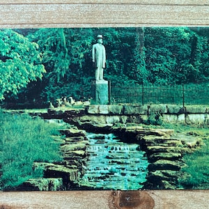 May include: A postcard featuring a statue of a man in a suit and hat standing on a stone pedestal. The statue is located in a park setting with a small waterfall and lush greenery.