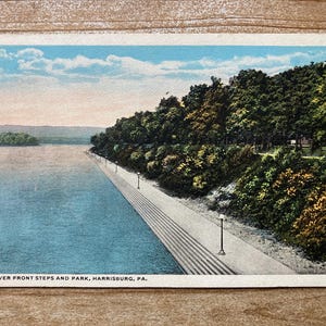 May include: A vintage postcard depicting a riverfront park with steps leading down to the water. The postcard is in color and shows a blue sky with white clouds, green trees, and a blue river. The text on the postcard reads "River Front Steps and Park, Harrisburg, PA."