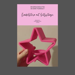Könnte beinhalten: Ein leuchtend rosa sternförmiges Objekt mit einer kleinen kreisförmigen Vertiefung in der Mitte. Das Objekt wird in einer Hand gehalten. Der Text auf dem rosa Hintergrund lautet "3D DRUCKDATEI IM 3MF FORMAT" und "Knödelstern mit Sitzstange".