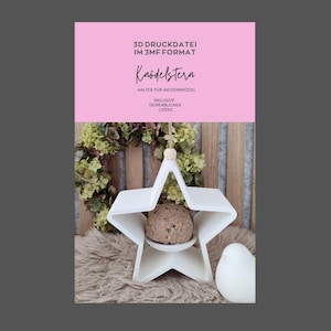 Könnte beinhalten: Ein weißer, sternförmiger Vogelfutterhalter mit einem runden Meisenknödel in der Mitte. Der Halter hängt an einer Schnur mit einer Holzperle. Im Hintergrund befindet sich ein rosa Schild mit deutschem Text und ein Kranz.