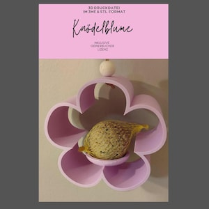 Könnte beinhalten: Ein hellrosa, blütenförmiges Vogelfutterhaus mit einem kleinen, runden Sitzplatz und einer Kugel aus Vogelfutter in einem Netz. Das Futterhaus trägt den Text "Knödelblume" und weitere Texte auf Deutsch.
