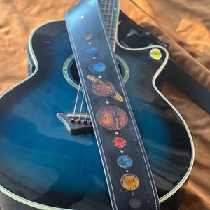 Może przedstawiać: Niebieska gitara akustyczna z czarnym sk&oacute;rzanym paskiem do gitary z wzorem planet w Układzie Słonecznym.