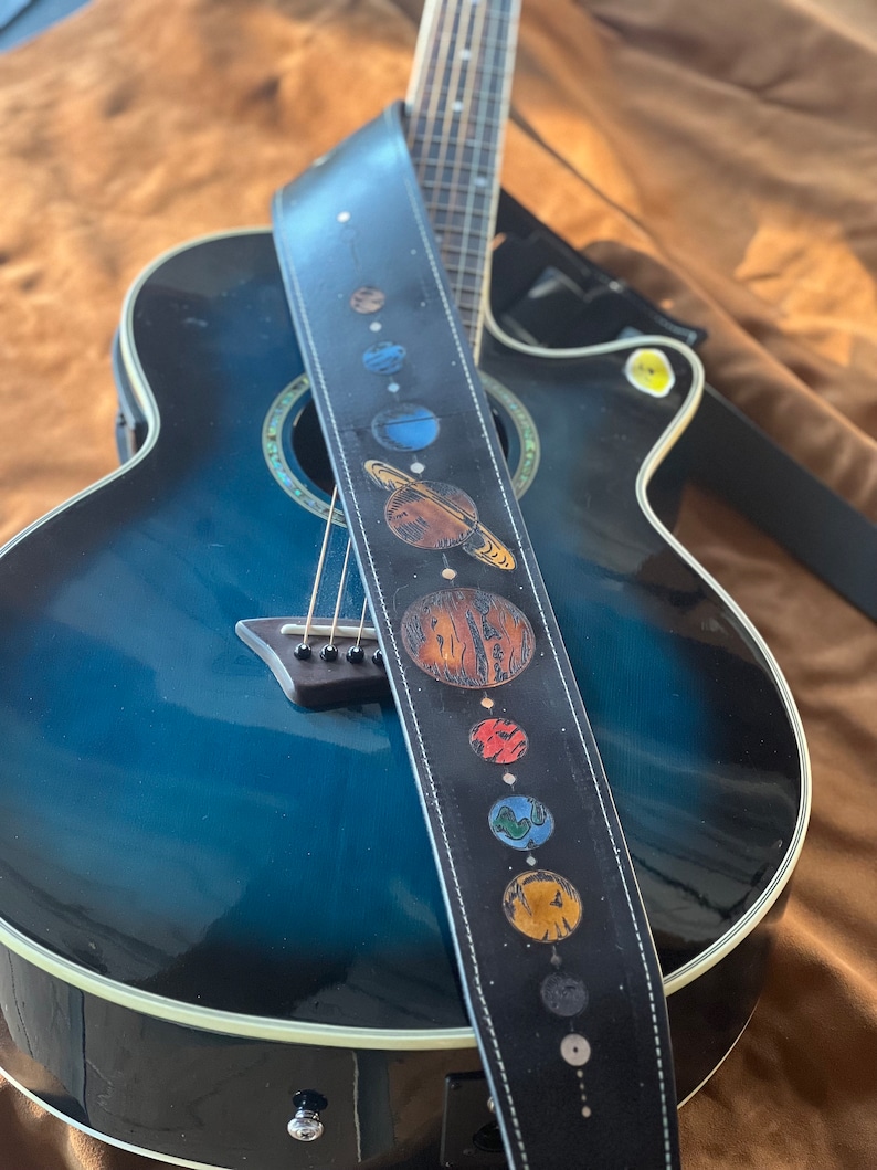 Może przedstawiać: Niebieska gitara akustyczna z czarnym sk&oacute;rzanym paskiem do gitary z wzorem planet w Układzie Słonecznym.