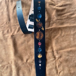 Może przedstawiać: Czarny sk&oacute;rzany pasek do gitary z wzorem układu słonecznego. Pasek ma czarne tło z linią planet w r&oacute;żnych kolorach, w tym czerwonym, pomarańczowym, ż&oacute;łtym, niebieskim i zielonym.