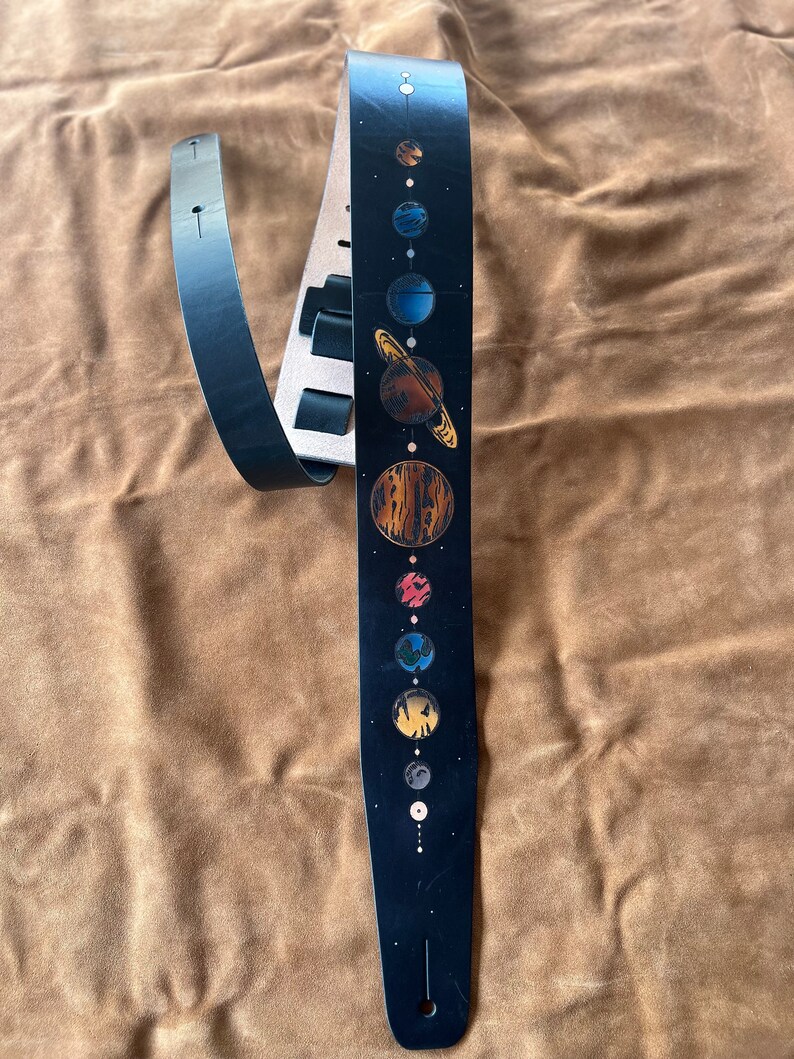 Może przedstawiać: Czarny sk&oacute;rzany pasek do gitary z wzorem układu słonecznego. Pasek ma czarne tło z linią planet w r&oacute;żnych kolorach, w tym czerwonym, pomarańczowym, ż&oacute;łtym, niebieskim i zielonym.