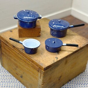 Könnte beinhalten: Ein Set aus vier Miniatur-Kochgeschirrteilen aus blauem Emaille mit weißen Sprenkelmustern. Die Sammlung umfasst zwei Töpfe mit Deckel und Seitengriffen, einen Topf mit Deckel und langem schwarzen Griff sowie eine offene Pfanne mit schwarzem Griff.
