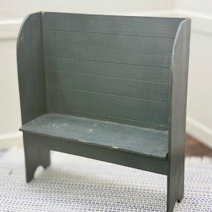 May include: A small, dark green wooden bench with a backrest. The bench has a simple design with a seat and a back made of horizontal planks. The legs are cut with a decorative curve. The bench is painted in a matte finish.