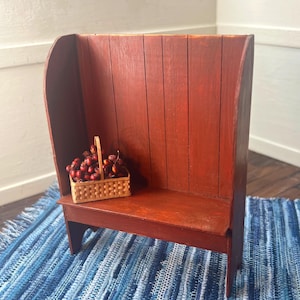 May include: A miniature red wooden bench with a high back and a small woven basket filled with red berries. The bench is placed on a blue and white striped rug, creating a rustic and charming scene.