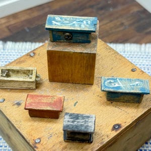 May include: A collection of miniature wooden furniture pieces, including a small chest of drawers, a rectangular box with handles, and several small tables. The pieces are painted in shades of blue, red, and grey, and are arranged on a wooden surface.