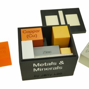 May include: A black box with a lid open, revealing a set of small cubes representing different metals and minerals. The cubes are labeled with their chemical symbols and names, including copper (Cu), zinc, silver (Ag), and potash (K20). The box is labeled "Metals & Minerals by Jerry Loo".