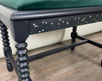 May include: A black painted stool with a dark green velvet cushion top. The stool has turned legs with a floral design painted on the top edge. The stool is sitting on a wood floor.