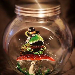 May include: A glass jar with a silver lid, painted with a green frog wearing a top hat and sitting on a red and white mushroom.
