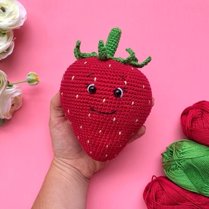 May include: A crocheted strawberry plush toy with a smiling face and green leaves. The toy is being held in a hand against a pink background.