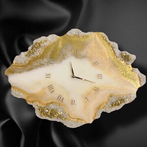 May include: A decorative wall clock with a unique, organic shape. The clock face is a creamy white with gold Roman numerals and hands. The edges are trimmed with gold and crystal-like accents, creating a luxurious look.