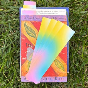 A bookmark that is a gradient of pink, blue and yellow pastel colors. This is a picture of a few of the bookmarks sitting on top of a pile of books such as a don miguel ruiz book.