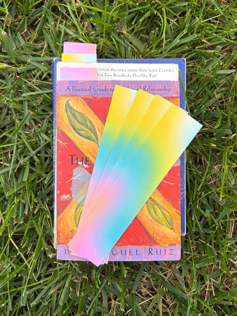 A bookmark that is a gradient of pink, blue and yellow pastel colors. This is a picture of a few of the bookmarks sitting on top of a pile of books such as a don miguel ruiz book.
